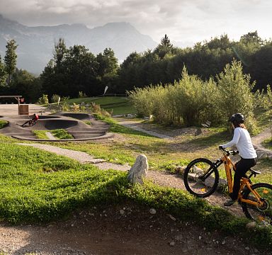 Ein Mountainbiker auf einem Pfad in einer grünen Landschaft, umgeben von Bäumen und Bergen im Hintergrund bei bewölktem Himmel. Ein Spielplatz ist ebenfalls sichtbar.