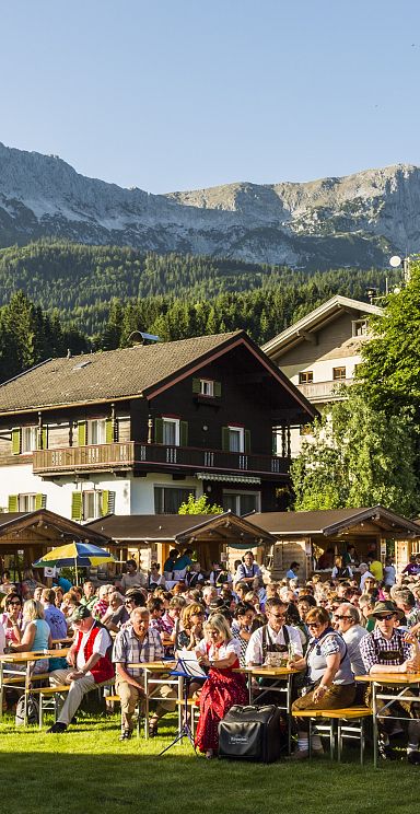 People sitting at picnic tables in a lush, green park with mountains in the background, enjoying a sunny day and festive atmosphere.