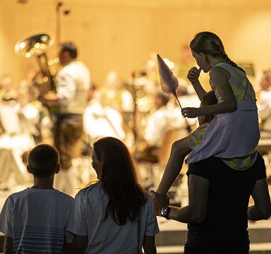 Ein Kind sitzt auf den Schultern eines Erwachsenen, während es Zuckerwatte isst. Im Hintergrund spielt ein Orchester auf einer Bühne. Abendstimmung.
