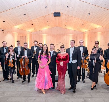 Eine Gruppe von Musikerinnen und Musikern in festlicher Kleidung, posierend mit ihren Instrumenten auf einer Bühne in einem holzgetäfelten Raum.