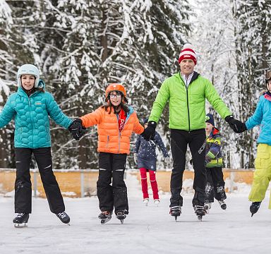 Familie mit Kindern beim Eislaufen im Freien, umgeben von verschneiten Bäumen. Alle tragen bunte Winterkleidung und scheinen Spaß zu haben.