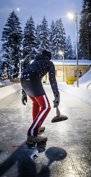 Person beim Eisstockschießen auf einem schneebedeckten Platz in winterlicher Umgebung, mit Lichtern und einer Holzhütte im Hintergrund.