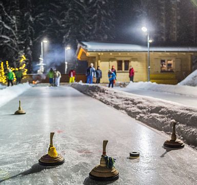 Mehrere Personen spielen Eisstockschießen auf einer beleuchteten Eisbahn neben einer gemütlichen Hütte im Schnee, umgeben von winterlicher Landschaft.