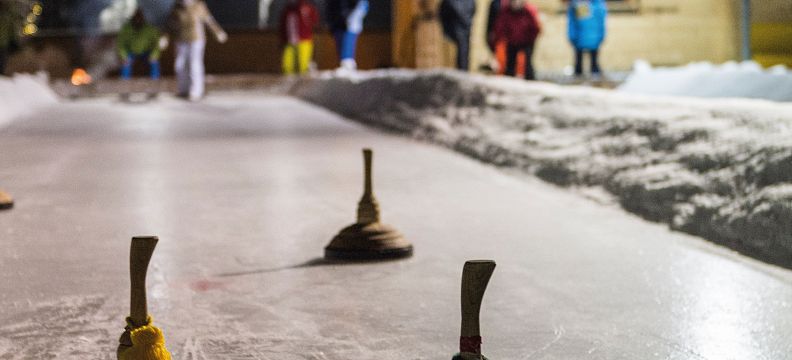 Gruppe von Menschen spielt Eisstockschießen auf einer beleuchteten Eisbahn; Vordergrund zeigt nahaufgenommene Eisstöcke.