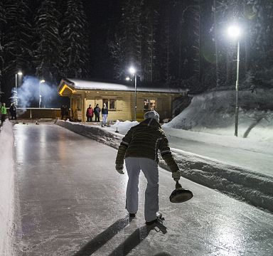 Person beim Eisstockschießen auf einer beleuchteten Eisbahn in einer verschneiten Winterlandschaft. Im Hintergrund eine Hütte mit weiteren Personen.