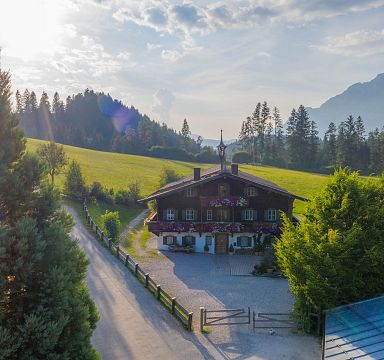 Idyllische Landschaft mit einem traditionellen alpinen Haus umgeben von grünen Wiesen und Bäumen, im Hintergrund sanfte Berge unter blauem Himmel.