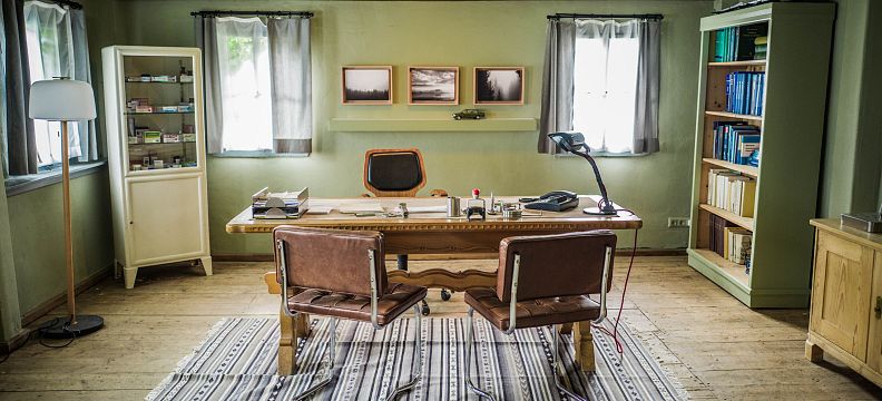Rustikales Arbeitszimmer mit Holzboden, großen Fenstern, einem Holztisch und zwei Ledersesseln. Bücherregale und Bilder schmücken den Raum.