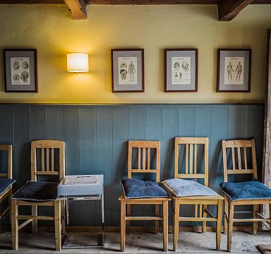 Holzstühle mit blauen Kissen stehen an einer grauen Wand in einem rustikalen Raum, dekoriert mit gerahmten Bildern und einer Holztür.