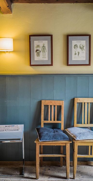 Holzstühle mit blauen Kissen stehen an einer grauen Wand in einem rustikalen Raum, dekoriert mit gerahmten Bildern und einer Holztür.