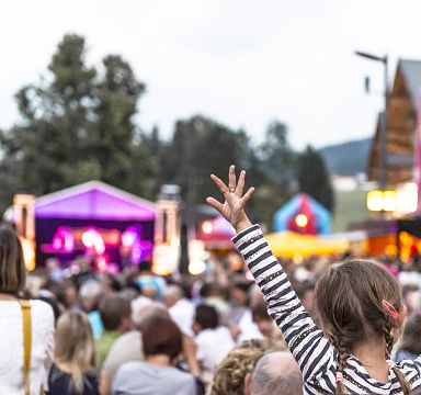 Outdoor concert scene with a large crowd and a young girl with raised arms enjoying the music. Colorful stage lights and tents add to the festive atmosphere.