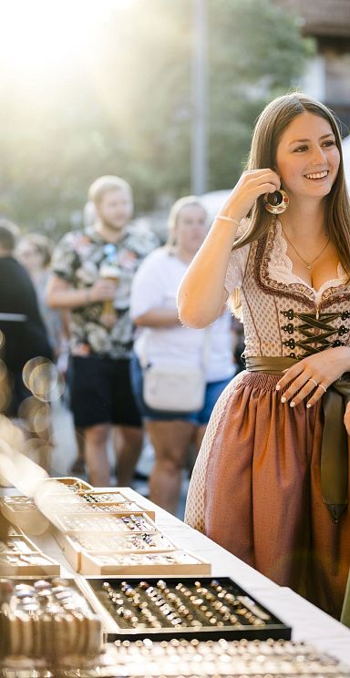 People in traditional attire visiting a market in the Wilder Kaiser region, with various stalls and a backdrop of mountains.