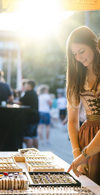 Zwei Frauen in traditionellen Trachten betrachten Schmuck an einem Marktstand im Freien bei Sonnenuntergang. Die Szene wirkt lebhaft und gemütlich.