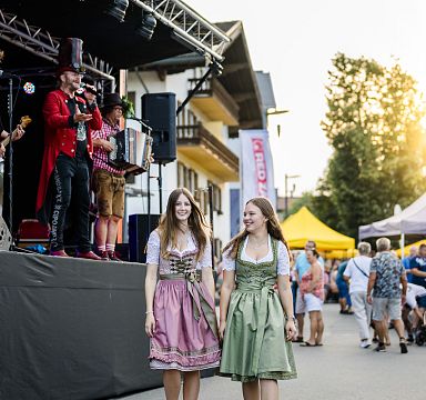 Zwei Frauen in traditionellen Dirndln gehen fröhlich an einer Bühne vorbei, wo Musiker auftreten. Menschen flanieren umher, während die Sonne scheint.