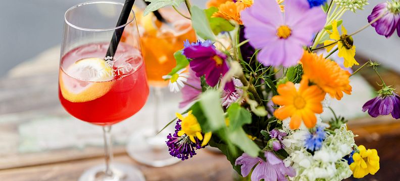 Zwei Aperol Spritz in Weingläsern mit Zitrone und Strohalmen, daneben ein bunter Blumenstrauß in einer Vase, auf einem Holztisch arrangiert.
