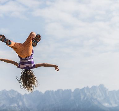 A person is captured mid-flip against the picturesque background of the Wilder Kaiser mountain range, under a mostly clear sky with scattered clouds.