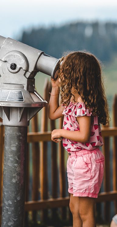 Two kids look through a telescope at the Wilder Kaiser mountains, standing by a wooden fence, enjoying the panoramic view.