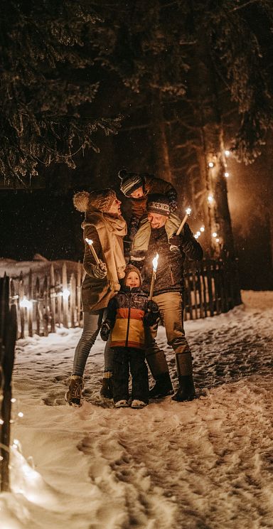 Eine Familie genießt einen winterlichen Spaziergang im Schnee. Sie tragen warme Kleidung und halten Fackeln, umgeben von Lichterketten in einer winterlichen Landschaft.