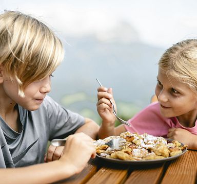 Zwei Kinder sitzen an einem Holztisch im Freien und essen gemeinsam ein Gericht, das wie Kaiserschmarrn aussieht. Sie wirken fröhlich und entspannt.