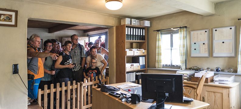 Eine Gruppe von Personen betrachtet interessiert etwas in einem Büro. Im Vordergrund steht ein Schreibtisch mit Computer, im Hintergrund sind Regale mit Akten.