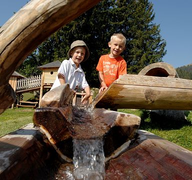 Zwei lachende Kinder spielen an einem Holzbrunnen in einer alpinen Wiese, umgeben von Bäumen und Bergen bei schönem Wetter.