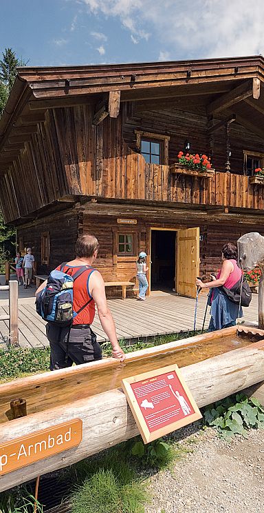 Holzhütte in alpiner Landschaft mit Personen davor, umgeben von Wald und Wiesen. Eine Kneipp-Anlage aus Holz befindet sich im Vordergrund.