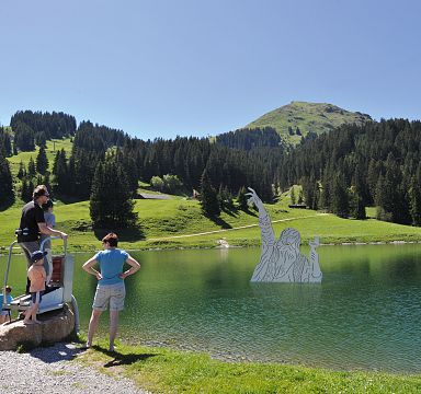 People enjoy a sunny day by a lake with green meadows and forests, and the Wilder Kaiser mountain in the background.
