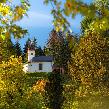 Eine malerische Kapelle steht auf einem Hügel, umgeben von bunten Herbstbäumen, unter klarem blauem Himmel. Die Szene strahlt Ruhe und Idylle aus.