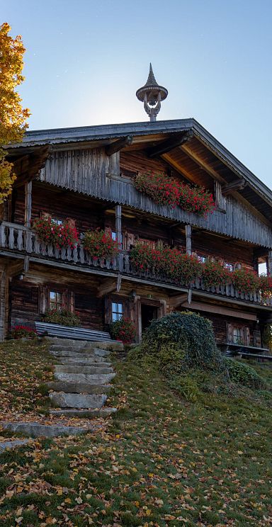 Alte Holzhütte in einer Herbstlandschaft, umgeben von buntem Laub und strahlendem Sonnenschein. Ein schöner Himmel und herbstliche Farbtöne dominieren die Szene.