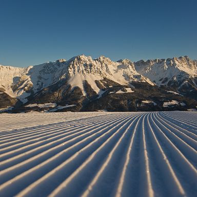 Schneebedeckte Bergkette im Hintergrund bei klarem Himmel, im Vordergrund perfekt präparierte Skipiste mit parallel verlaufenden Linien, Sonnenlicht taucht alles in ein sanftes Licht.