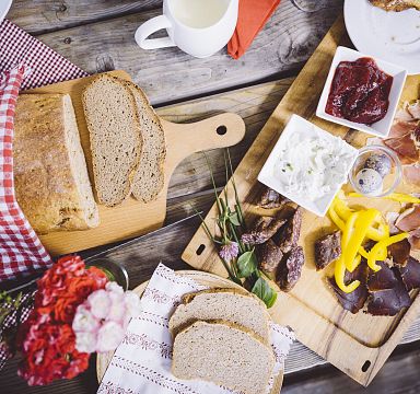 Rustikale Brotzeit mit Schinken, Brot, Gemüse und Dips auf Holztisch. Einladend angerichtet mit karierten Servietten und gemütlicher Atmosphäre.