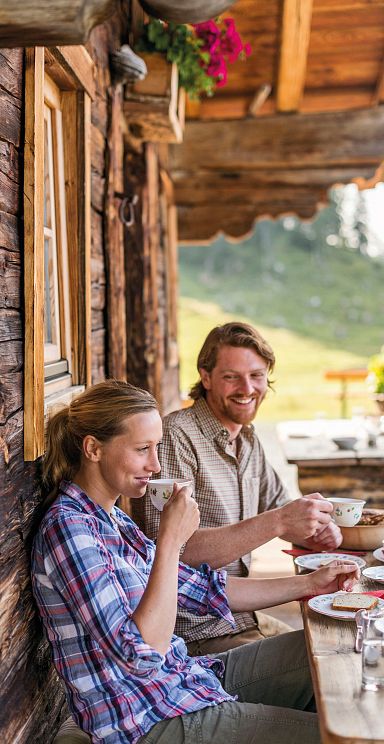 Zwei Personen sitzen auf einer Berghütte aus Holz und genießen Kaffee. Im Hintergrund eine grüne Berglandschaft und ein Tisch mit Blumen.
