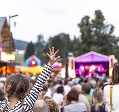 An outdoor concert scene at Wilder Kaiser, with a child in the foreground raising arms excitedly. Colorful lights illuminate the stage, surrounded by a lively crowd.