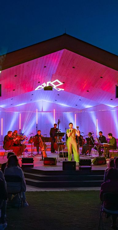 Open-Air-Konzert in einem Pavillon mit bunten Lichtern. Musiker auf der Bühne spielen vor einem Publikum, während der Abendhimmel im Hintergrund zu sehen ist.