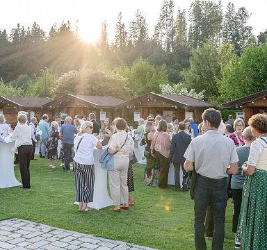 Eine Gruppe von Menschen versammelt sich bei Sonnenuntergang auf einer grünen Wiese vor Holzhütten, wahrscheinlich bei einer Veranstaltung im Freien.