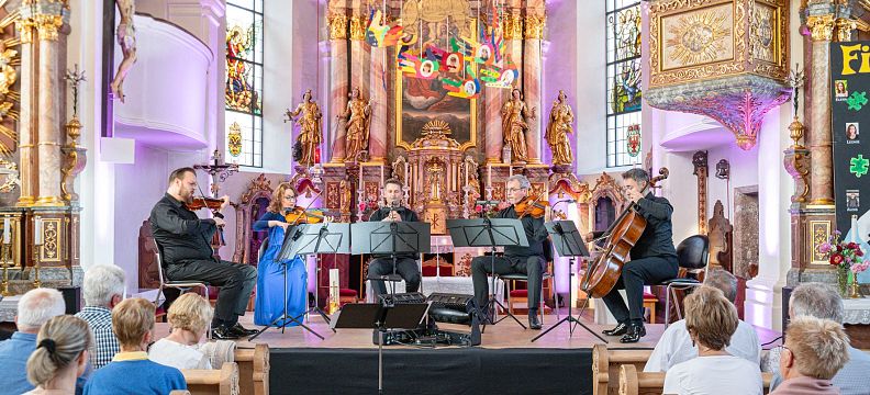 Kammermusikensemble spielt in einer barocken Kirche vor Publikum. Die Kirche ist prachtvoll dekoriert mit Statuen und einem kunstvollen Altar.