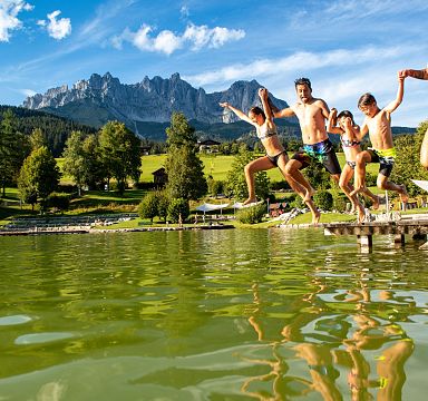 People jumping into a lake with the Wilder Kaiser mountains in the background, enjoying a sunny day.
