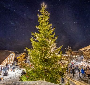 Christmas market at night with a large decorated tree and festive lights, set against the backdrop of snowy Wilder Kaiser mountains.