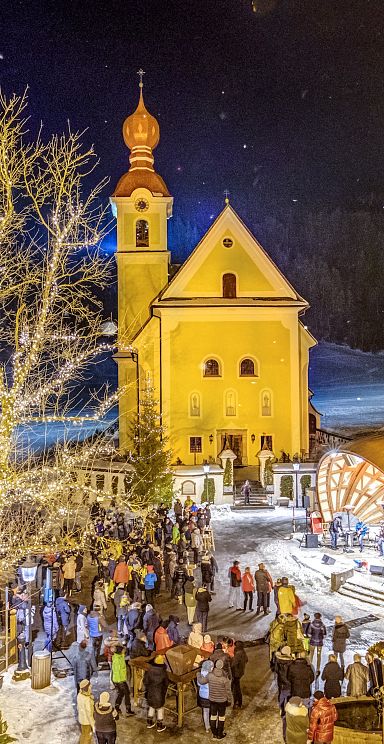 Illuminated church with snowy surroundings and people gathering in Wilder Kaiser at night.