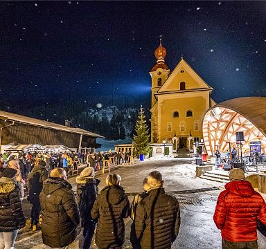 A snowy evening at Wilder Kaiser, with people in warm clothes gathered around a church, the scene is illuminated by festive lights and a starry sky.