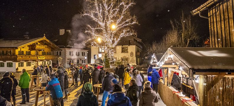 People at a winter market in Wilder Kaiser, surrounded by festive stalls and a lit tree. Snow is falling, adding to the charming alpine village atmosphere.