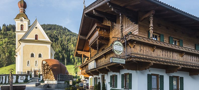 Bayerisches Dorf mit traditionellem Holzhaus, daneben eine Kirche mit Zwiebelturm und grünen Bergen im Hintergrund, unter einem blauen Himmel.