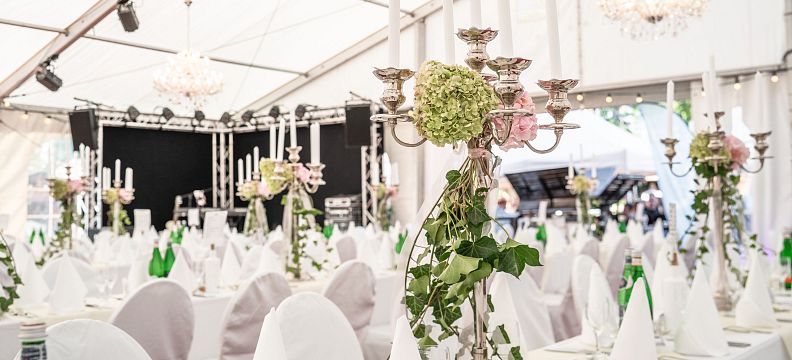 Elegant event setup with white tablecloths, decorated with floral centerpieces and candles in a spacious venue. Tables are neatly arranged with chairs.