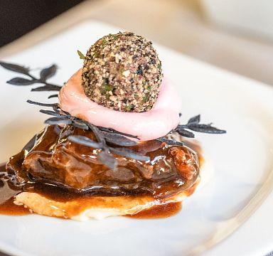 A gourmet dish featuring a piece of meat topped with pink sauce and a herb-crusted ball, garnished with leaves, on a white plate.