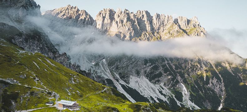 Berglandschaft mit grünen Wiesen und einer Hütte im Vordergrund, im Hintergrund ragen felsige Gipfel empor, während Wolken sanft darüber hinweg ziehen.