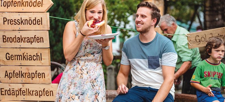 Two people enjoying traditional Austrian food at an outdoor event, with wooden signs indicating various local dishes like "Speckknödel" and "Käsespätzle."