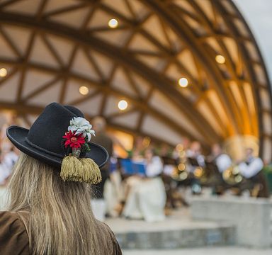 Person in traditioneller Tracht mit Hut und Blumen schaut auf ein Freiluftkonzert. Die Musiker tragen ebenfalls Trachten. Die Bühne hat eine Holzstruktur.