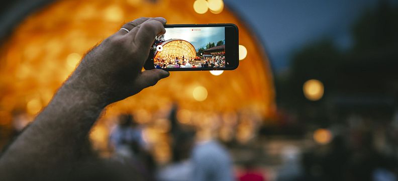 Person hält ein Smartphone und fotografiert eine beleuchtete, halbrunde Bühne bei Nacht, während im Vordergrund Menschen verschwommen sichtbar sind.