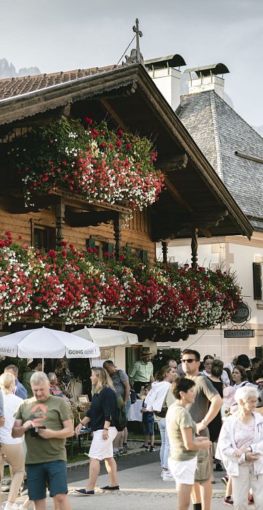 Alpine village scene with a traditional house, vibrant flowers, and people enjoying the day. Wilder Kaiser mountains in the background under a clear sky.
