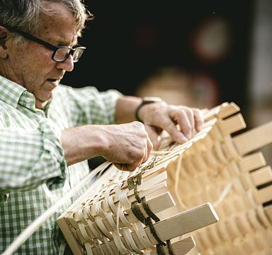 Ein älterer Mann mit Brille und kariertem Hemd flechtet konzentriert einen Korb aus hellem Holzstreifen. Die Umgebung ist unscharf, der Fokus liegt auf der Handarbeit.