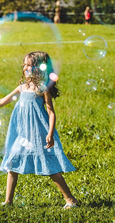 Ein Kind in einem blauen Sommerkleid spielt auf einer grünen Wiese mit großen Seifenblasen unter strahlendem Sonnenschein.